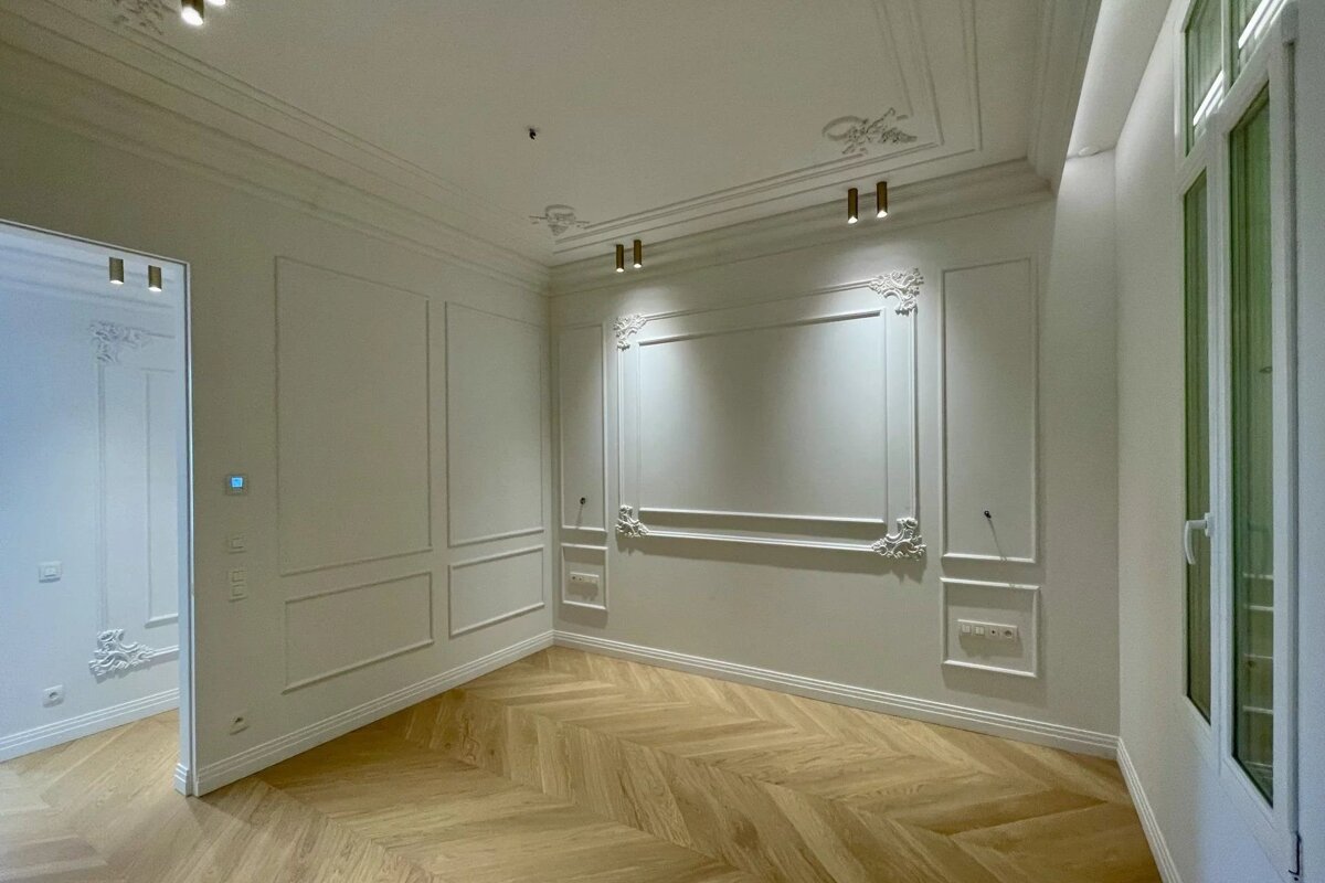 Empty room with elegant white walls, ornate molding, herringbone wood floor, and modern lights. A window is on the right.