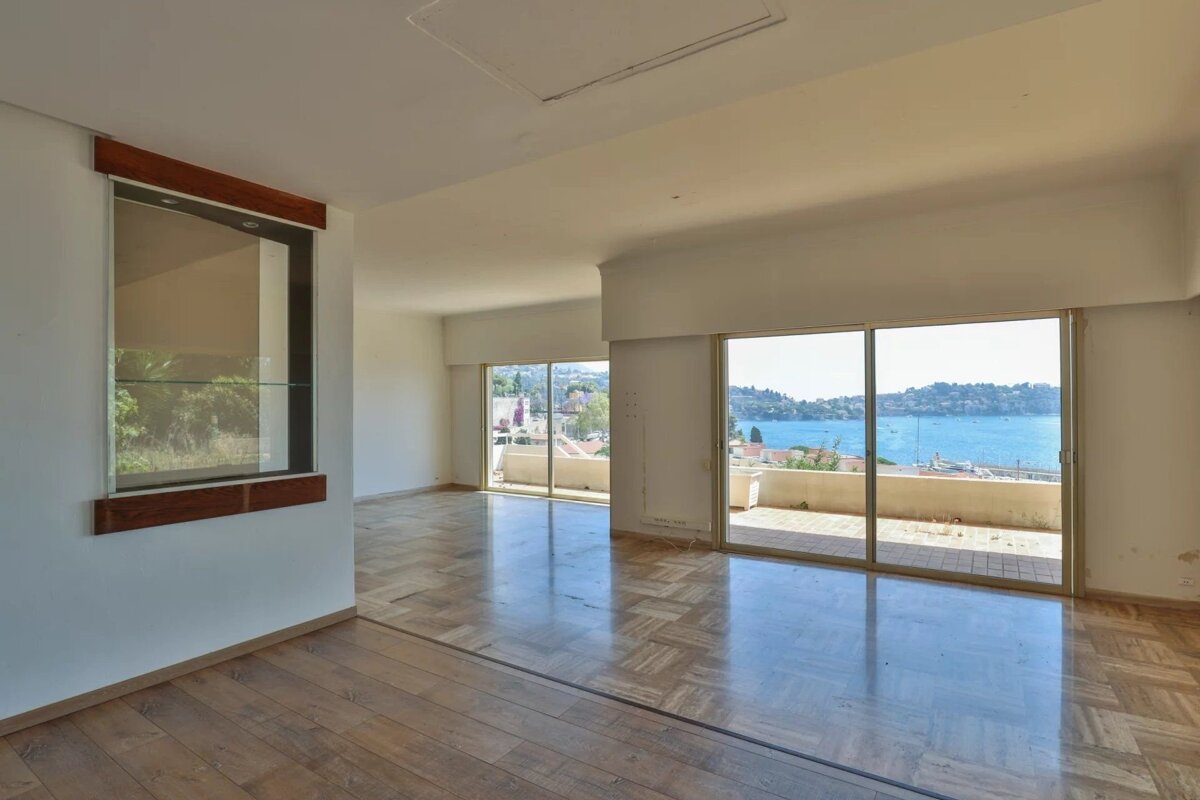 An empty room with glossy parquet floors, a built-in display, and large sliding glass doors opening to a balcony with a stunning sea view.