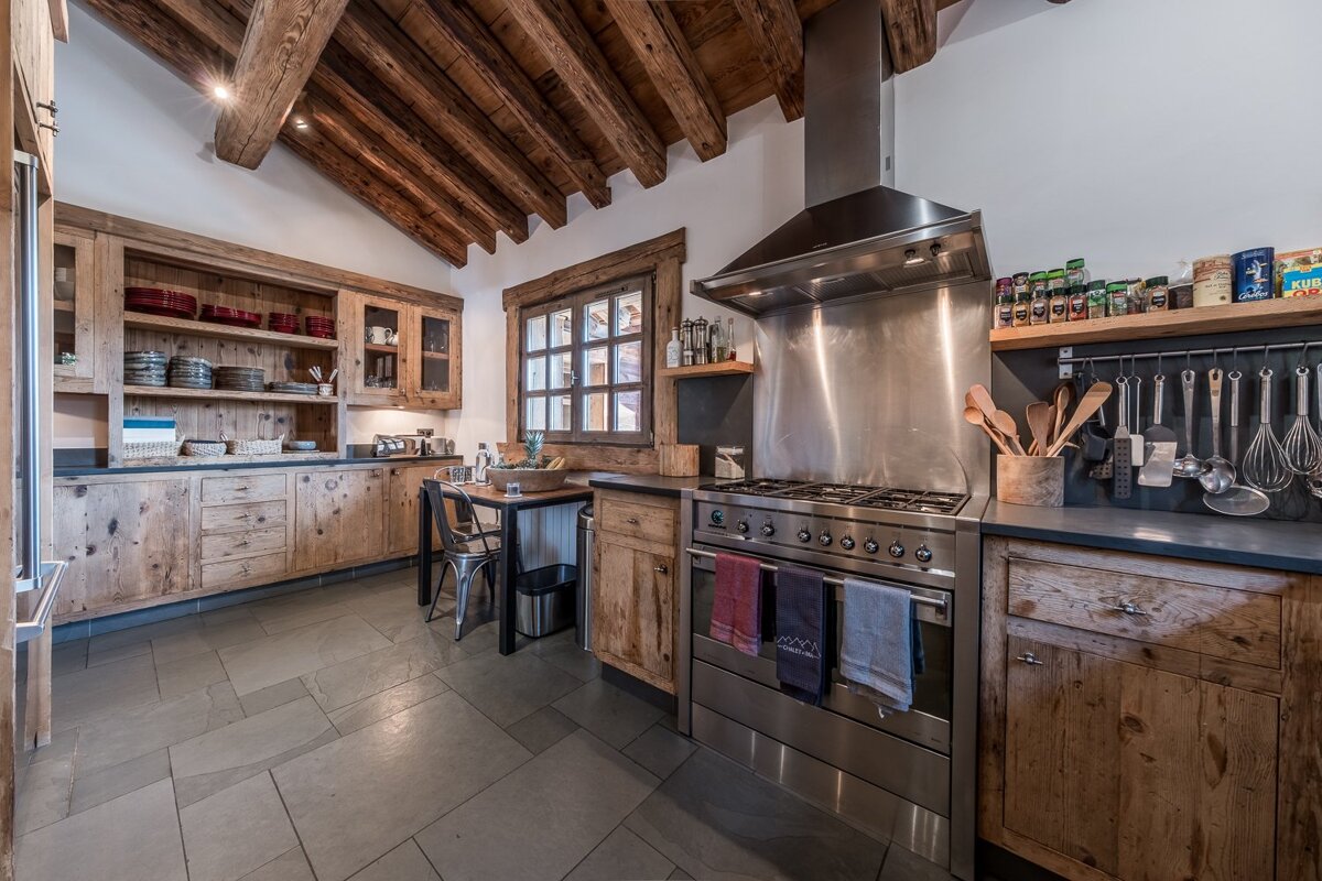Cozy, rustic kitchen featuring exposed wooden beams, distressed wood cabinetry, stone floor, and modern stainless steel appliances.