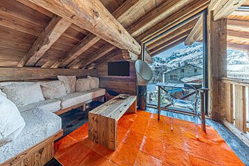 Cozy wooden attic living room in an alpine chalet with a bright orange rug and a large window overlooking snowy mountains.