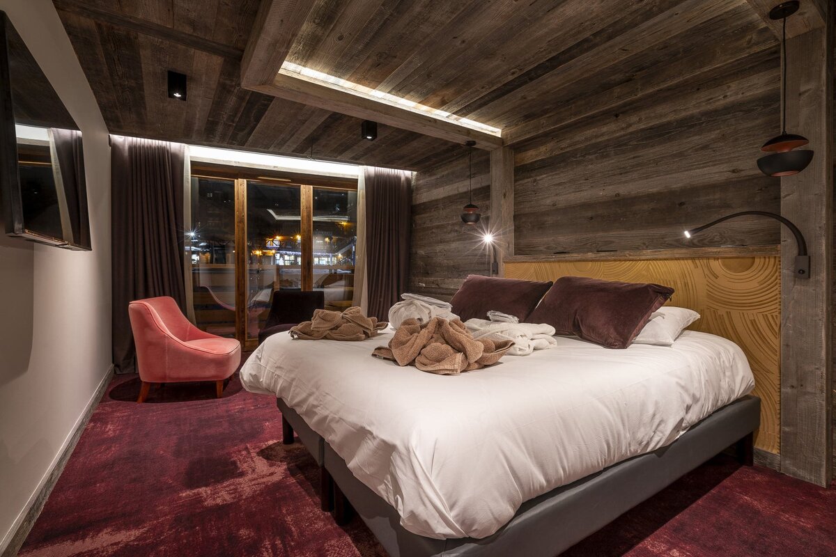 A cozy bedroom with rustic wood walls/ceiling, a large bed with robes, pink armchair, and a window looking out at a night scene.