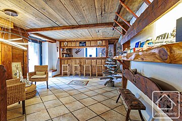 A rustic, wood-paneled interior features a bar with stools, tiled floor, wicker chairs, and exposed wooden beams. A modern light fixture hangs from the ceiling.