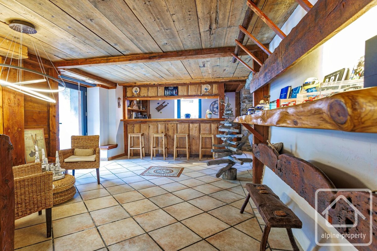 Cozy chalet interior with a rustic wooden bar, stools, wicker chairs, tiled floor, and wood-paneled ceiling. Modern and traditional elements blend.