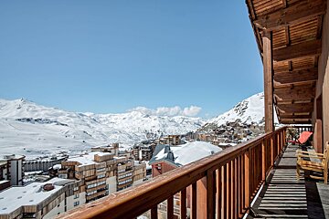Wooden balcony overlooking a snow-covered alpine ski resort and mountains under a clear blue sky.