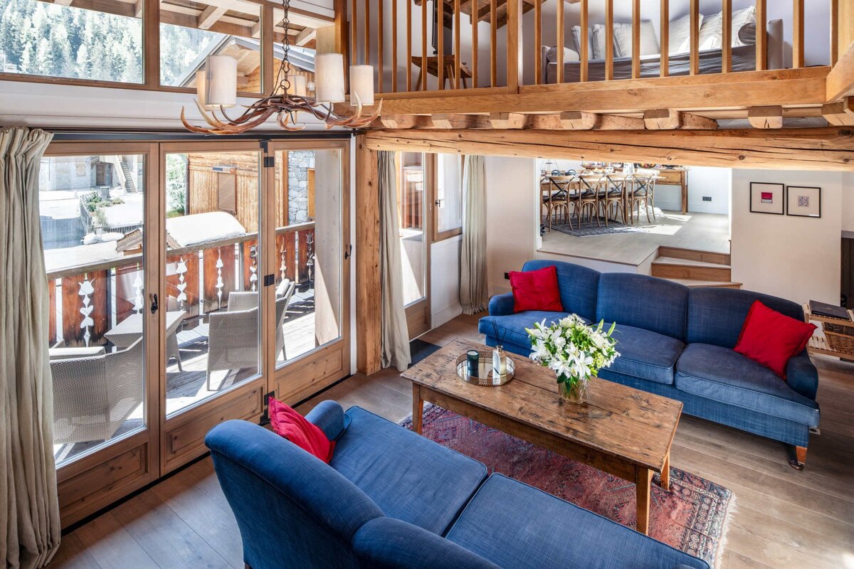 Cozy rustic chalet living room with blue sofas, a wooden coffee table, and large glass doors leading to a snowy balcony.
