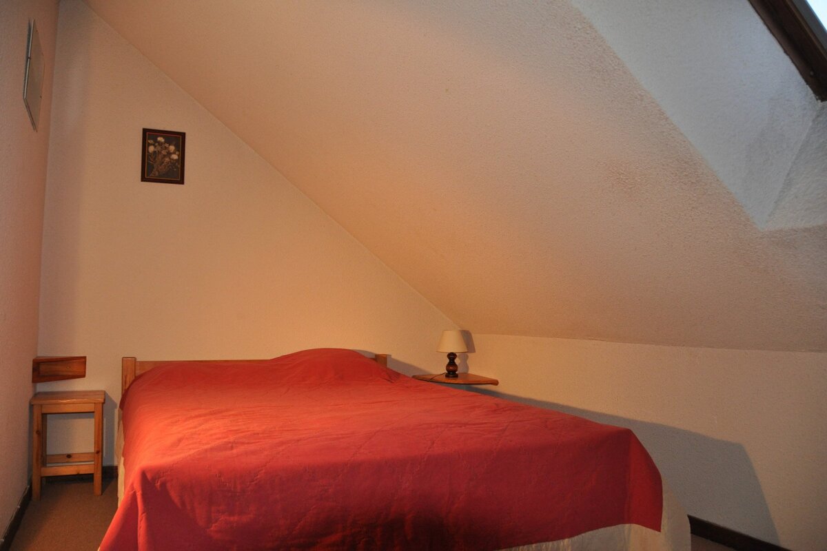 A cozy attic bedroom with a bed featuring a red comforter, a small bedside table with a lamp, and a picture on the slanted wall.