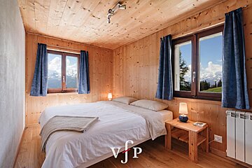 Cozy wooden bedroom with a double bed, warm lamps, and two windows showing scenic mountain views through blue curtains.