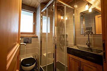 A modern bathroom with gray tiled walls, wooden ceiling, and a bright window. It features a toilet, a glass shower, and a stone sink on a wooden vanity.