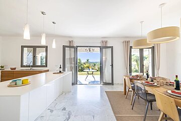 A modern, bright open-plan kitchen and dining area with white decor, marble floor, and large glass doors leading to a sunny garden patio.