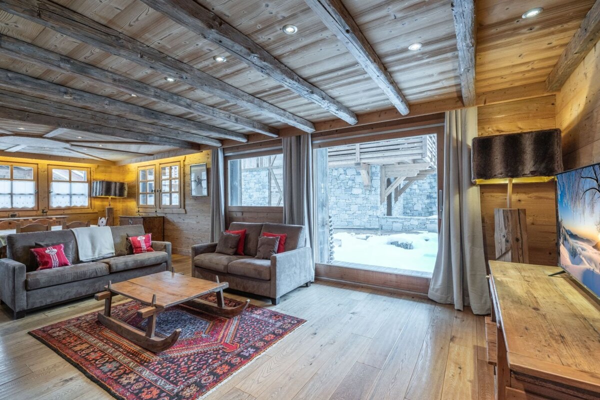 Cozy wooden chalet living room with two sofas, a sled-style coffee table on a patterned rug, and a snowy view through large windows.
