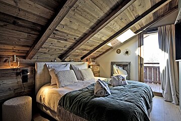 A warm, inviting attic bedroom featuring exposed wooden beams, a plush bed with a green blanket, a large mirror, and a door to a sunlit balcony.