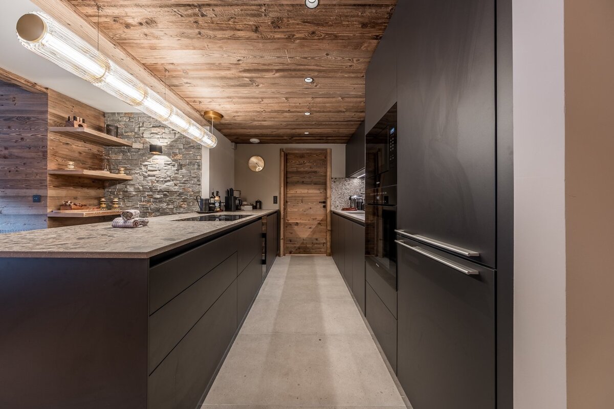 Modern kitchen with sleek dark cabinets, rustic wood ceiling & walls, and a stone accent wall. Features a large island and a long linear light.