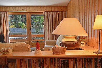 A lamp sits on a wooden table next to a bookshelf filled with books including one titled