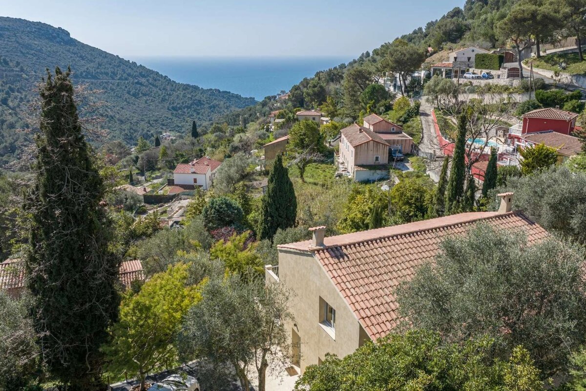 Hillside village with terracotta-roofed houses and lush trees, including cypress, cascading down to a vibrant blue ocean under a clear sky.