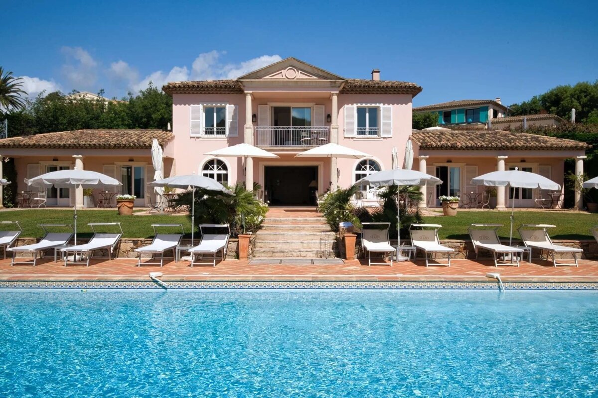 An elegant pink villa with a large blue swimming pool, sun loungers, and umbrellas, set against a clear blue sky.