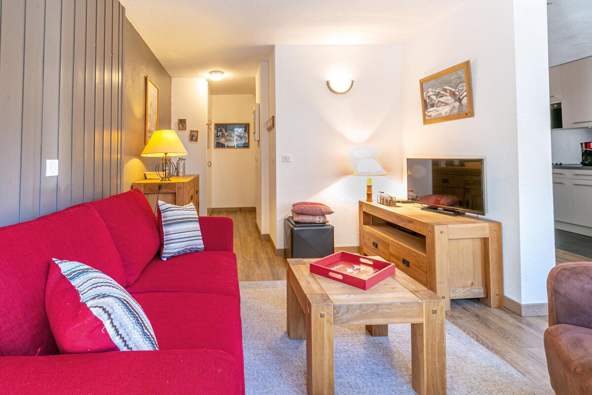 A cozy living room features a bright red sofa, wooden coffee table, and TV stand. Lamps cast a warm glow, and a hallway leads to a glimpse of the kitchen.