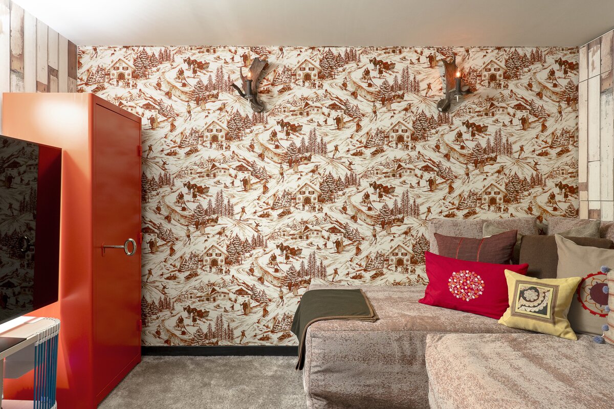 Cozy room with brown & white winter ski chalet wallpaper, a red cabinet, plush sofa with colorful cushions, and antler sconces.