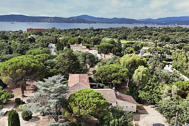 An aerial view shows a lush, tree-filled landscape with houses, a bay dotted with boats, and distant mountains under a cloudy sky.
