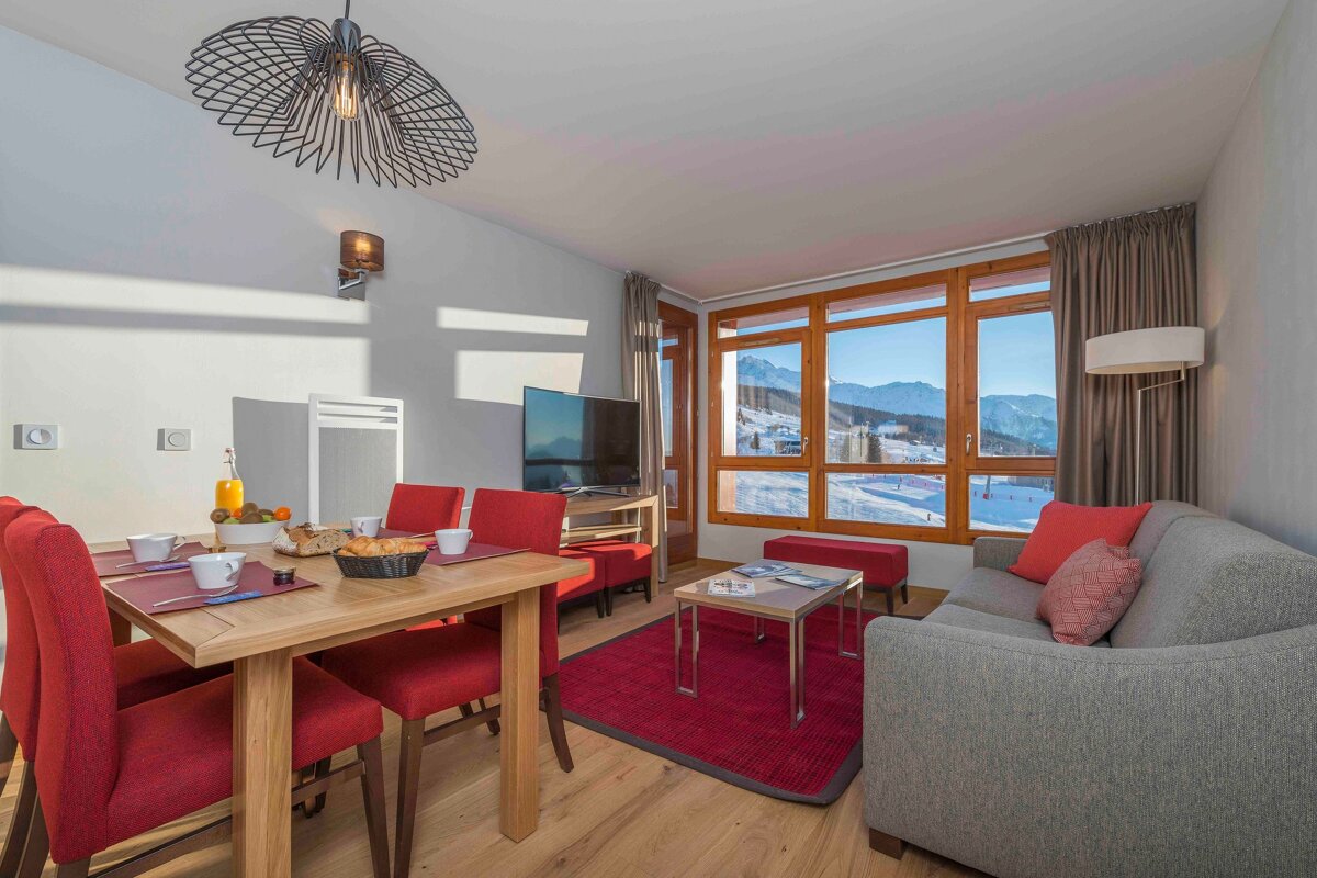 A modern living/dining room with breakfast set, red chairs/rug, and a stunning snowy mountain/ski slope view from large windows.