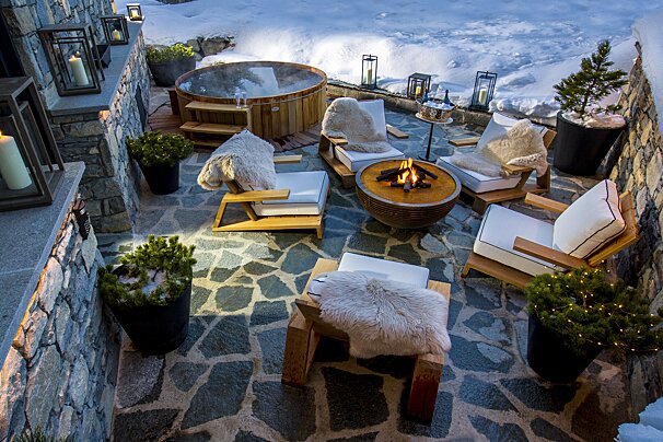 An inviting winter patio features a hot tub, glowing fire pit, comfy chairs with fur blankets, and atmospheric lanterns, set against a snowy backdrop.