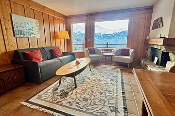 Cozy wood-paneled living room in a chalet with a grey sofa, fireplace, and large windows looking out at snowy mountains.