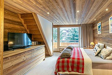 A cozy, wood-paneled chalet bedroom with a large bed, red plaid throw, TV under stairs, and a window offering a snowy mountain view.