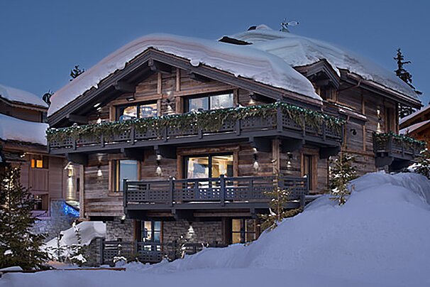 A rustic wooden chalet covered in fresh snow, with festive lights adorning its balconies, under a twilight sky.