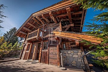 A rustic wooden chalet with a prominent balcony and stone base, featuring exposed beams. It sits on a wooden deck surrounded by trees under a clear blue sky.