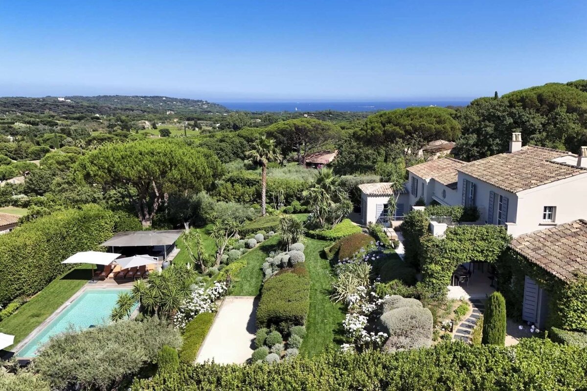 Aerial view of a beautiful villa with a pool and manicured gardens, nestled in a green landscape with a distant ocean view.