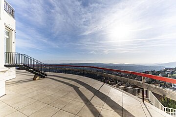 A spacious tiled terrace with railings offers a panoramic view of a valley and distant hills under a bright, sunny sky.