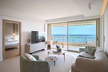 A chic, modern living room overlooks the ocean from a balcony, featuring contemporary furnishings and a connected bedroom visible through a doorway.