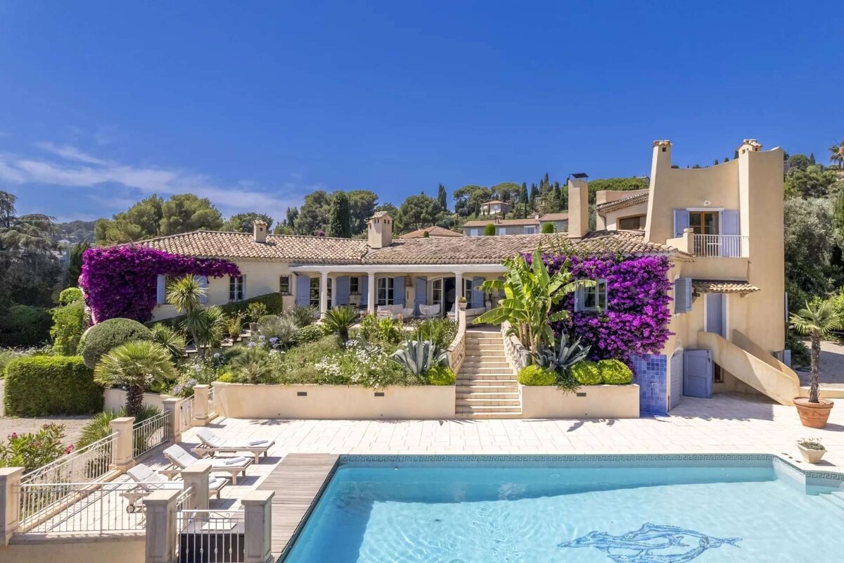A luxurious Mediterranean villa with vibrant purple bougainvillea, lush gardens, and a sparkling swimming pool under a clear blue sky.
