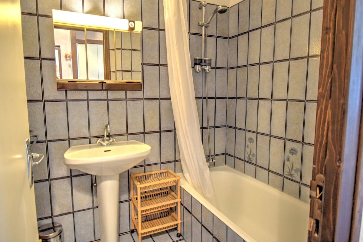A small bathroom with light tiled walls, featuring a white pedestal sink, a bathtub with shower, mirror, and a small wooden shelving unit.