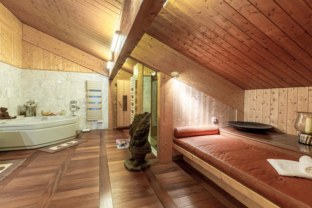 A cozy, rustic spa room with wood paneling, a large bathtub, sauna door, and a brown leather relaxation bench under an angled ceiling.