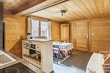 A rustic chalet-style room with wood-paneled walls/ceiling, a kitchen corner, dining table with chairs, a window, and a wooden door. Cozy and inviting.