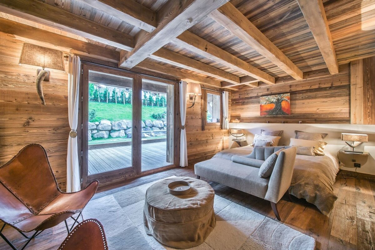 A luxurious, wood-paneled bedroom with exposed beams, a large bed, chaise lounge, and glass doors revealing a grassy outdoor area.