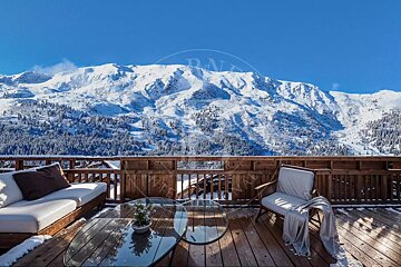 A stunning snowy mountain vista under a clear blue sky, viewed from a wooden deck with outdoor furniture lightly dusted with snow.