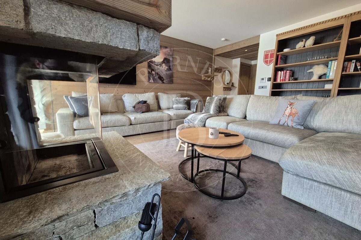 Cozy chalet living room with a stone fireplace, large grey sectional sofas, wooden nesting coffee tables, and a bookshelf.