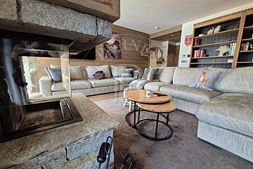 Cozy chalet living room with a stone fireplace, large grey sectional sofas, wooden nesting coffee tables, and a bookshelf.