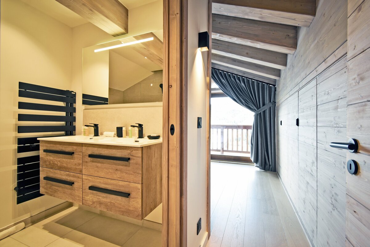 Modern chalet interior showing a wooden bathroom vanity on the left and a wood-paneled hallway leading to a balcony on the right.