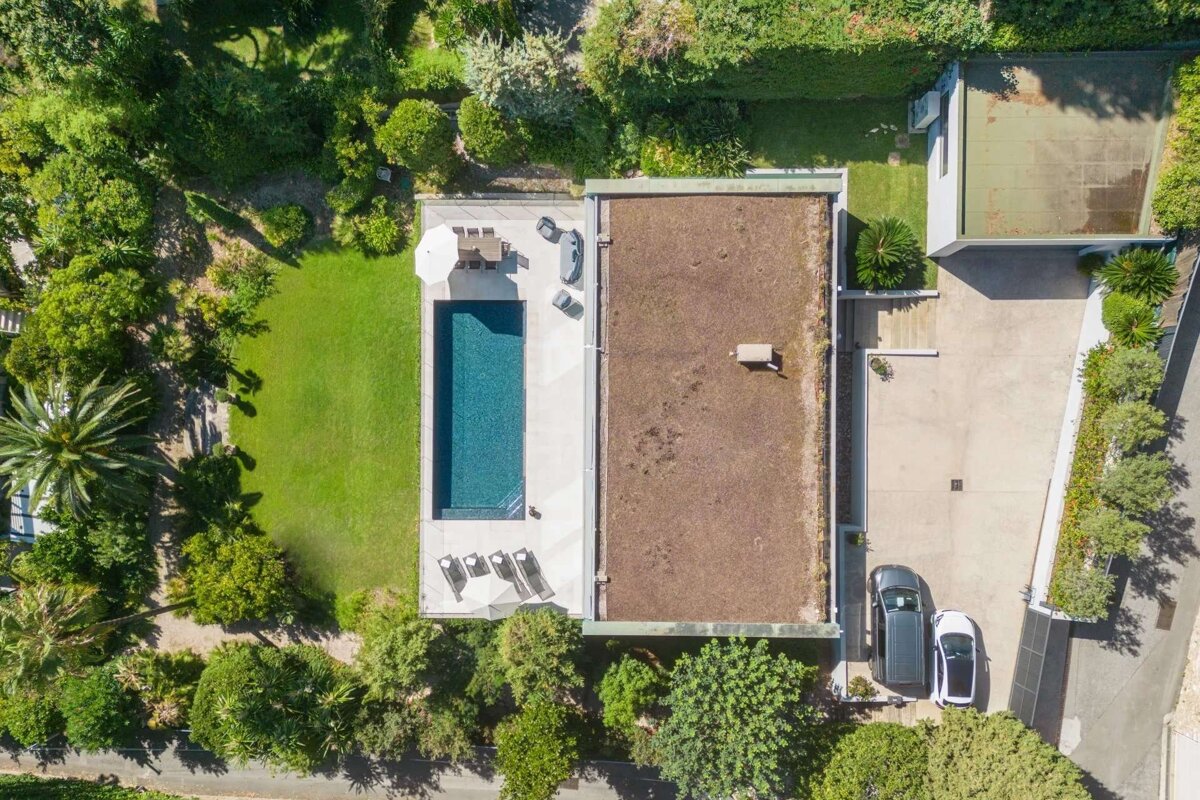 Aerial view of a modern house with a flat roof, a long swimming pool, green lawn, lush garden, and a driveway with two cars.