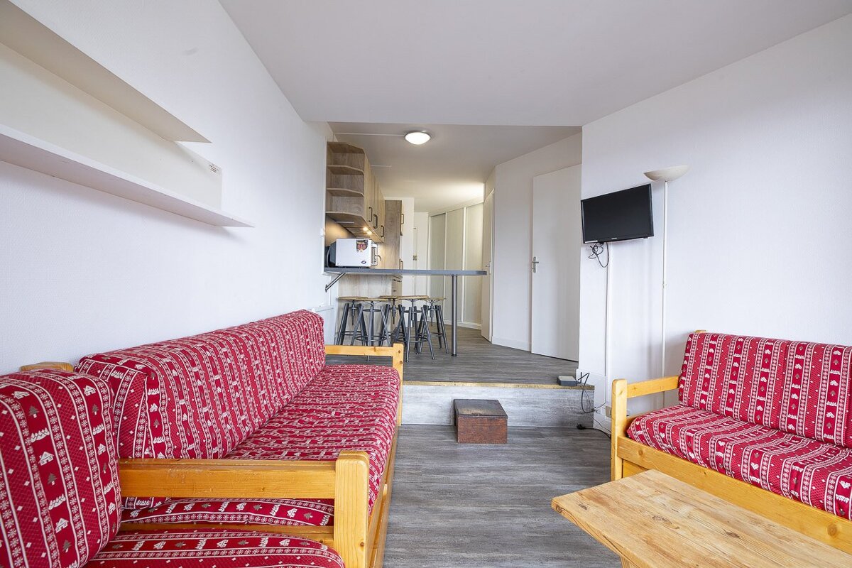 A bright living area with two red patterned sofas, white walls, and a raised kitchen with a bar & stools. Wood-effect flooring and a wall-mounted TV.