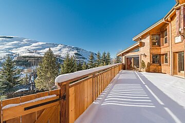 Snow-covered wooden chalet balcony overlooking pine trees and snow-capped mountains under a clear blue sky.