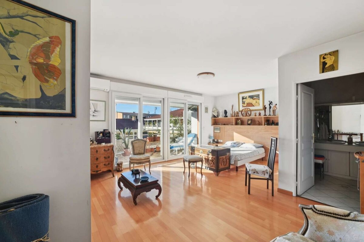 Sunlit room with wooden floors, balcony access, a bed, coffee table, chairs, and eclectic decor. An open door reveals another room.