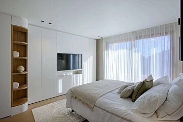 A bright, modern white bedroom featuring an integrated TV in sleek cabinetry, a bed with soft linens, and sheer curtains covering a large window.