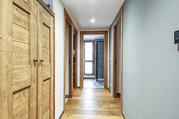 A bright hallway with light wooden floors, door frames, and a large wooden closet on the left. White and light gray walls lead to a bright window at the end.