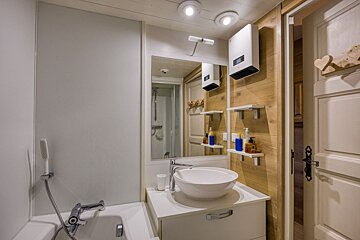 A cozy, modern bathroom featuring a tub, vessel sink vanity, mirror, wooden accent wall, wall heater, and toiletries on shelves.