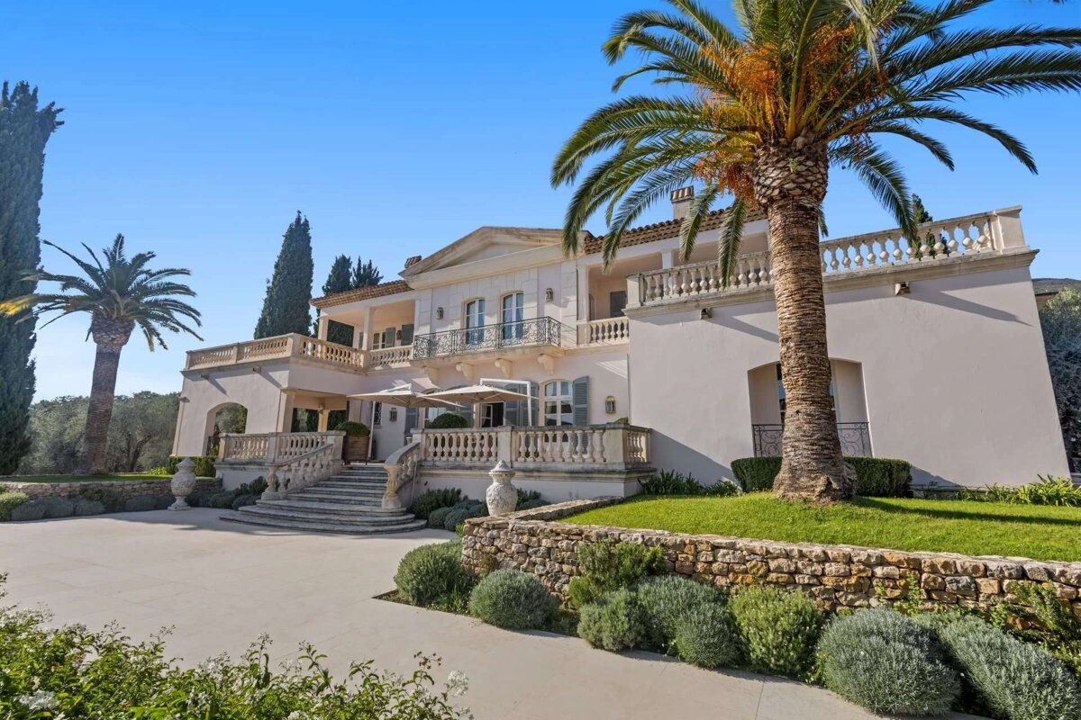 A grand, elegant villa with ornate stairs and balconies, flanked by lush palm trees and cypress trees, under a clear blue sky.