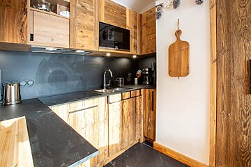 A modern rustic kitchen with warm knotty wood cabinets, dark countertops, and integrated appliances. A cutting board hangs on the wall, lit by warm spotlights.