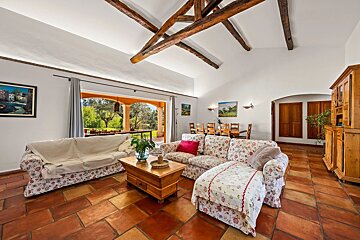 Rustic living room with terracotta tile floors, exposed wooden ceiling beams, floral patterned sofas, and open doors leading to a patio.
